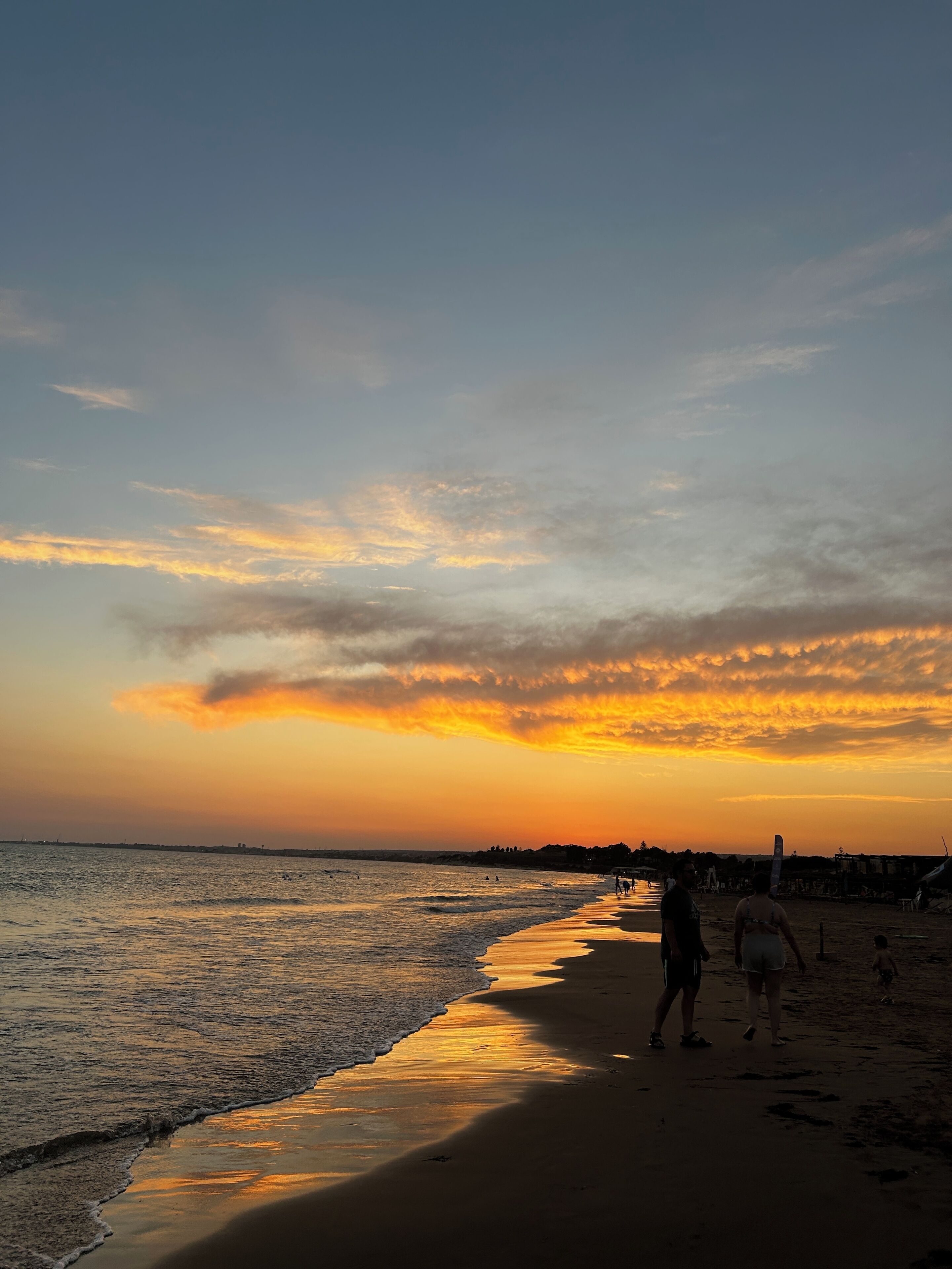 Une plage où des gens se promènent le long du rivage, un coucher de soleil en arrière-plan et des vagues calmes sur la mer.