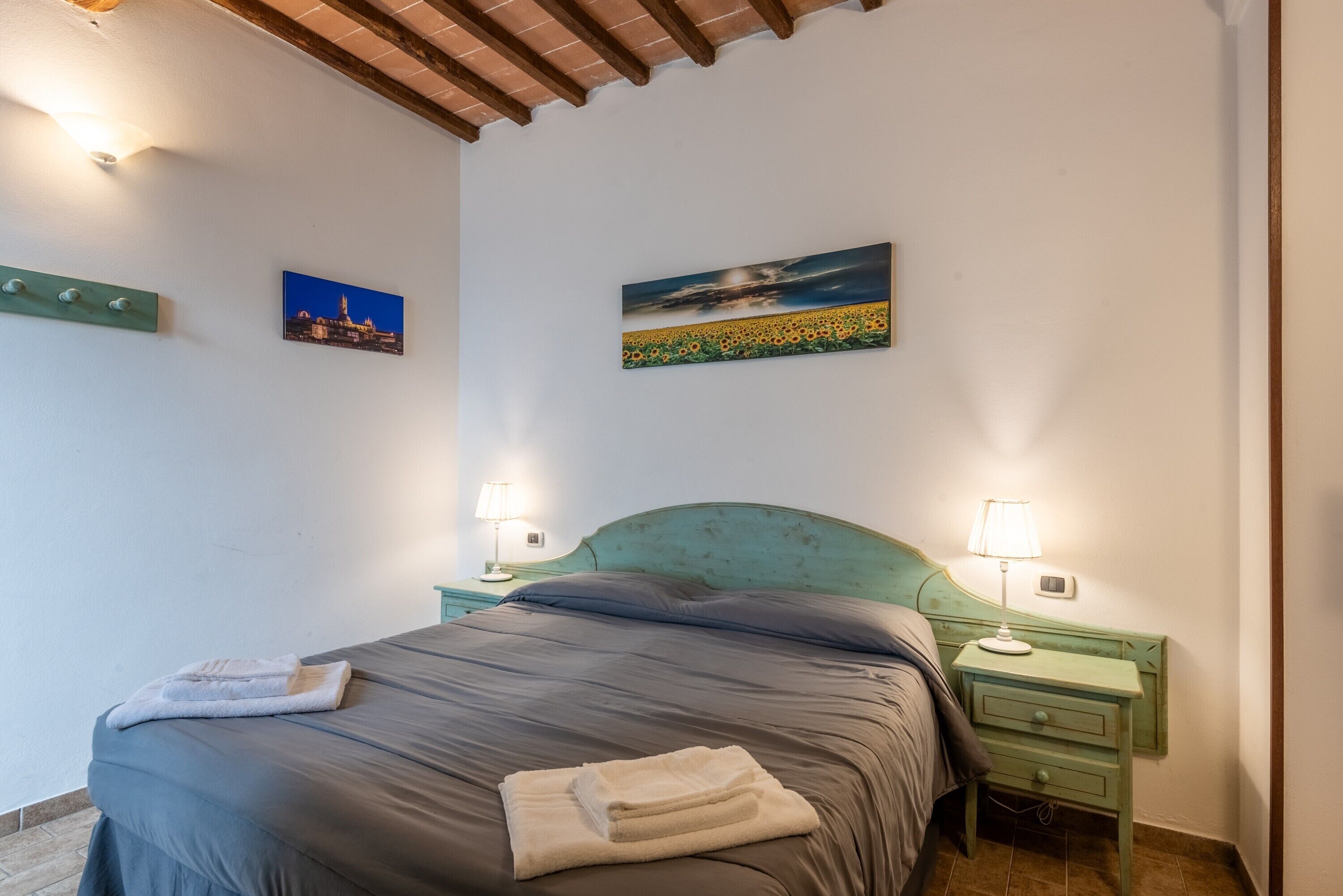 Une chambre à coucher avec un lit, deux tables de chevet avec des lampes, un tableau représentant un coucher de soleil sur un champ, et une petite table de nuit avec une lampe.