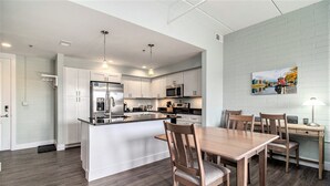 A modern kitchen with white cabinets, a wooden dining table, and a painting on the wall. - 2 Bedroom Downtown Waterfront Condo (Manistee)