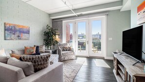 A modern living room with a grey sofa, a large TV, and a view of the city through the sliding glass doors. - 2 Bedroom Downtown Waterfront Condo (Manistee)