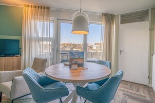 A round table with six chairs, a television on the left, and a view of houses through large windows.