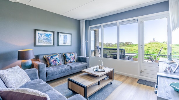 A modern living room with a grey sofa, a coffee table, and a large window offering a view of the outdoors. - 4 Person Apartment With sea View (De Koog)