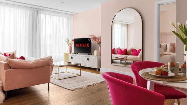 Ett modernt vardagsrum med en rosa soffa, ett matbord med rosa stolar, en tv på ett vitt skåp och en stor spegel på väggen.