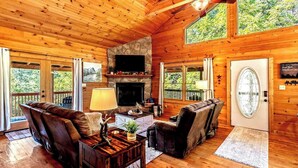 A cozy cabin interior with a fireplace, wooden furniture, and large windows offering a view of the forest. - New Listing! Mountain Views! 3B/3Ba Sleep 6-7, Hot Tub, Close to Everything! (Sevierville)