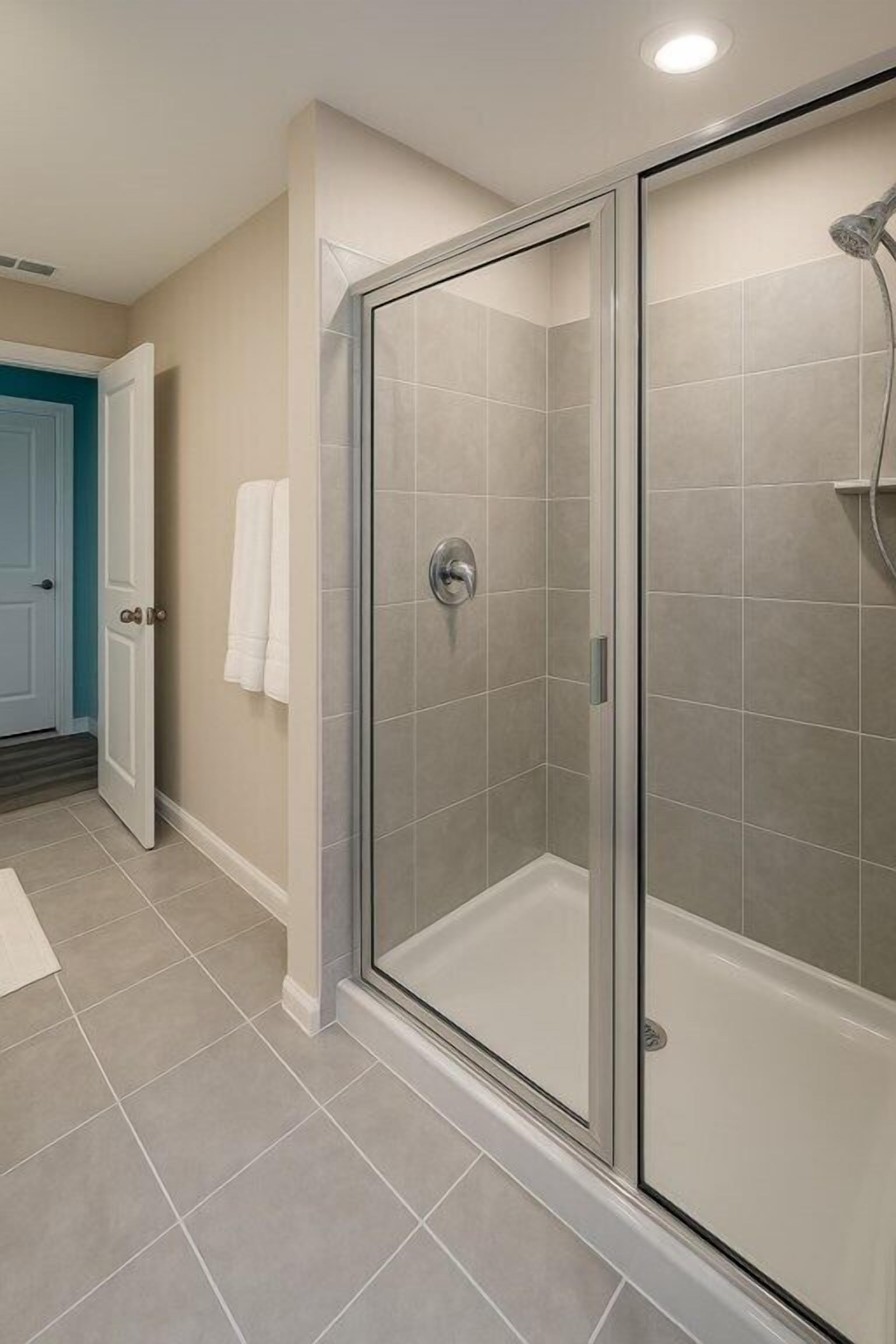 A glass-enclosed shower with a handheld showerhead, beige walls, and a white door in the background.