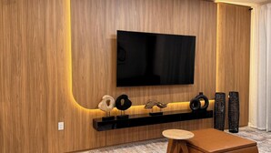 Una sala de estar moderna con un panel de madera, un televisor montado, un estante con adornos y un otomano de cuero marrón.