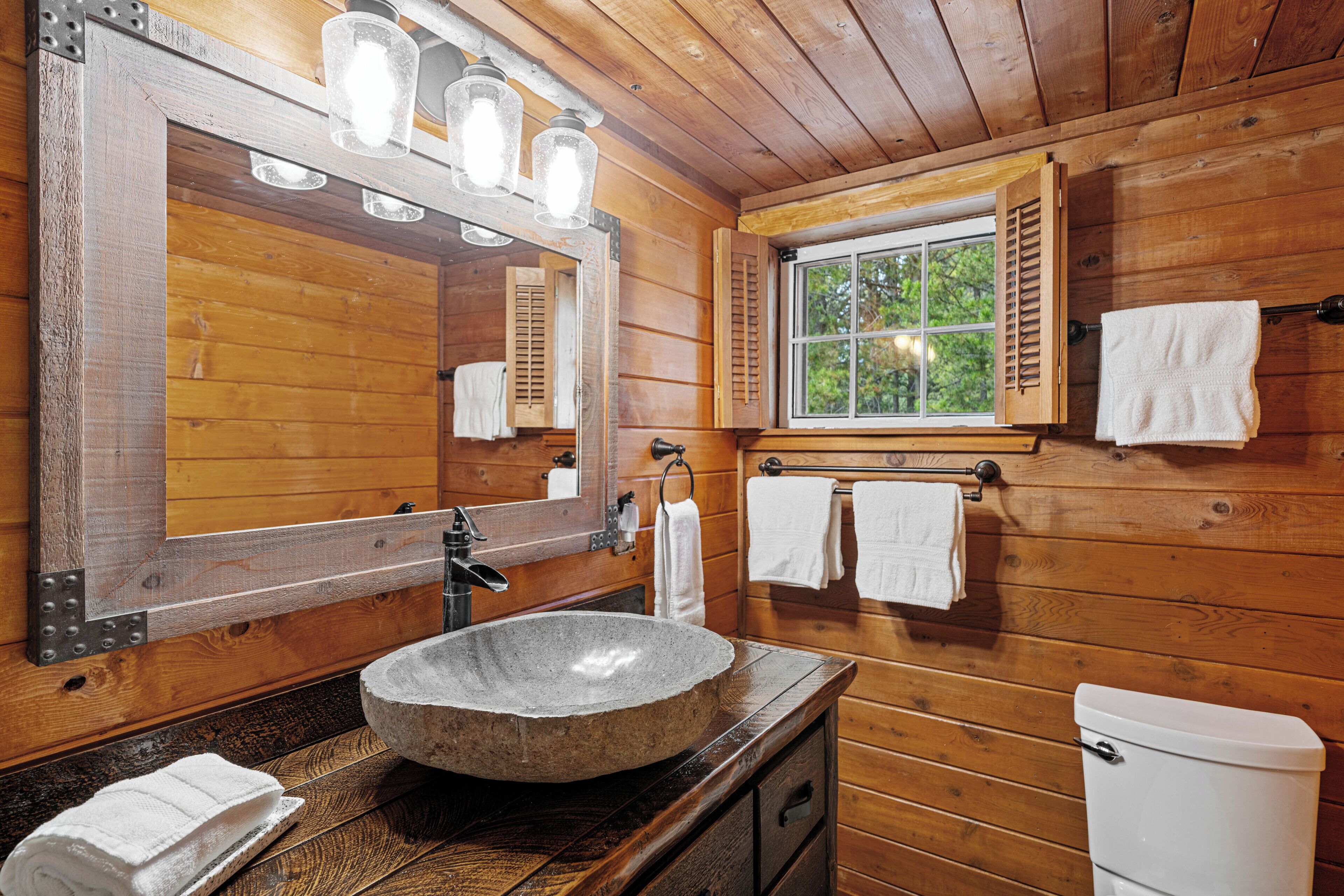 A wooden cabin bathroom with a stone sink, wooden cabinets, a large mirror, and a window with shutters.