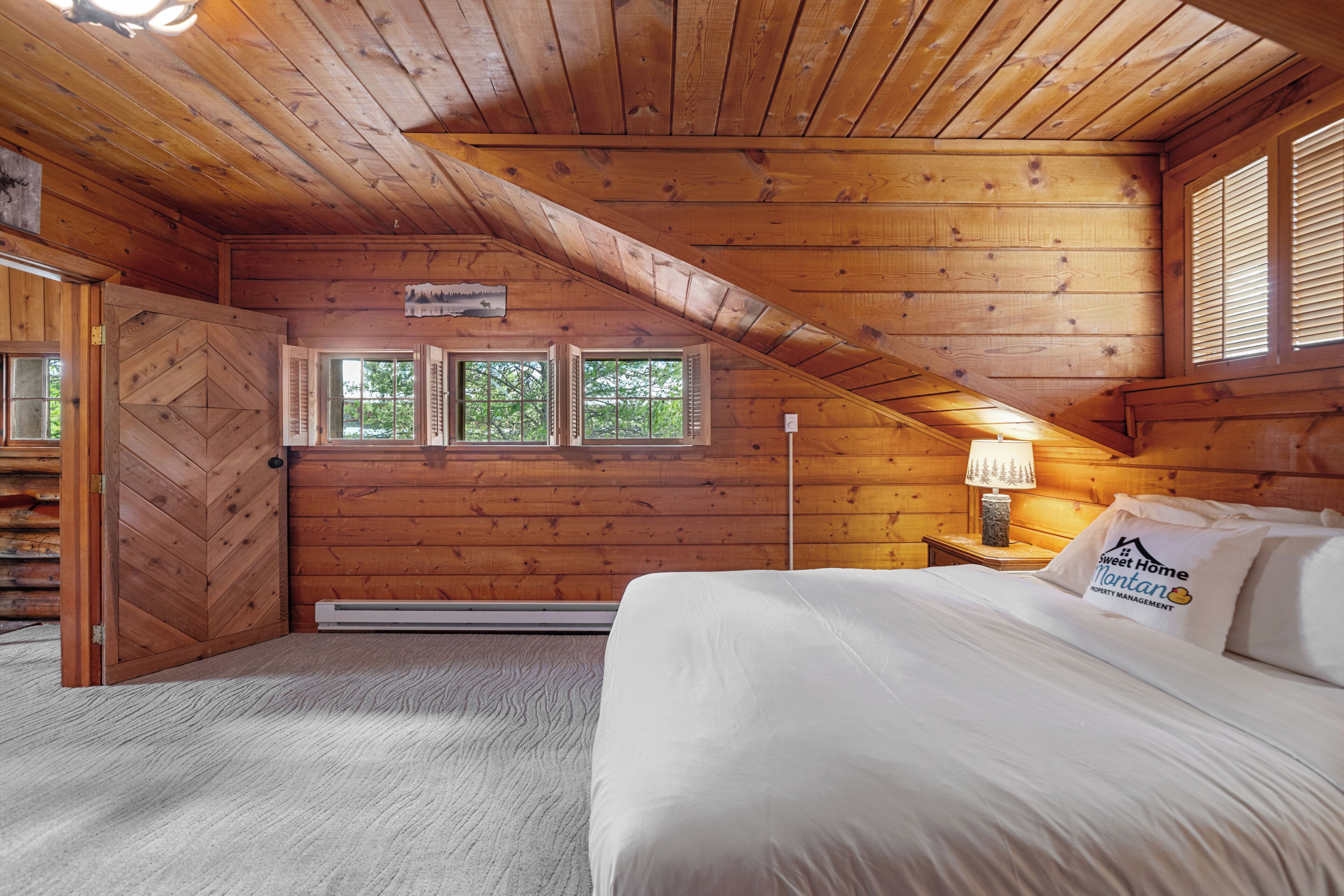 Un dormitorio de cabaña de madera con una cama, una mesita de noche, una lámpara y una ventana con contraventanas.