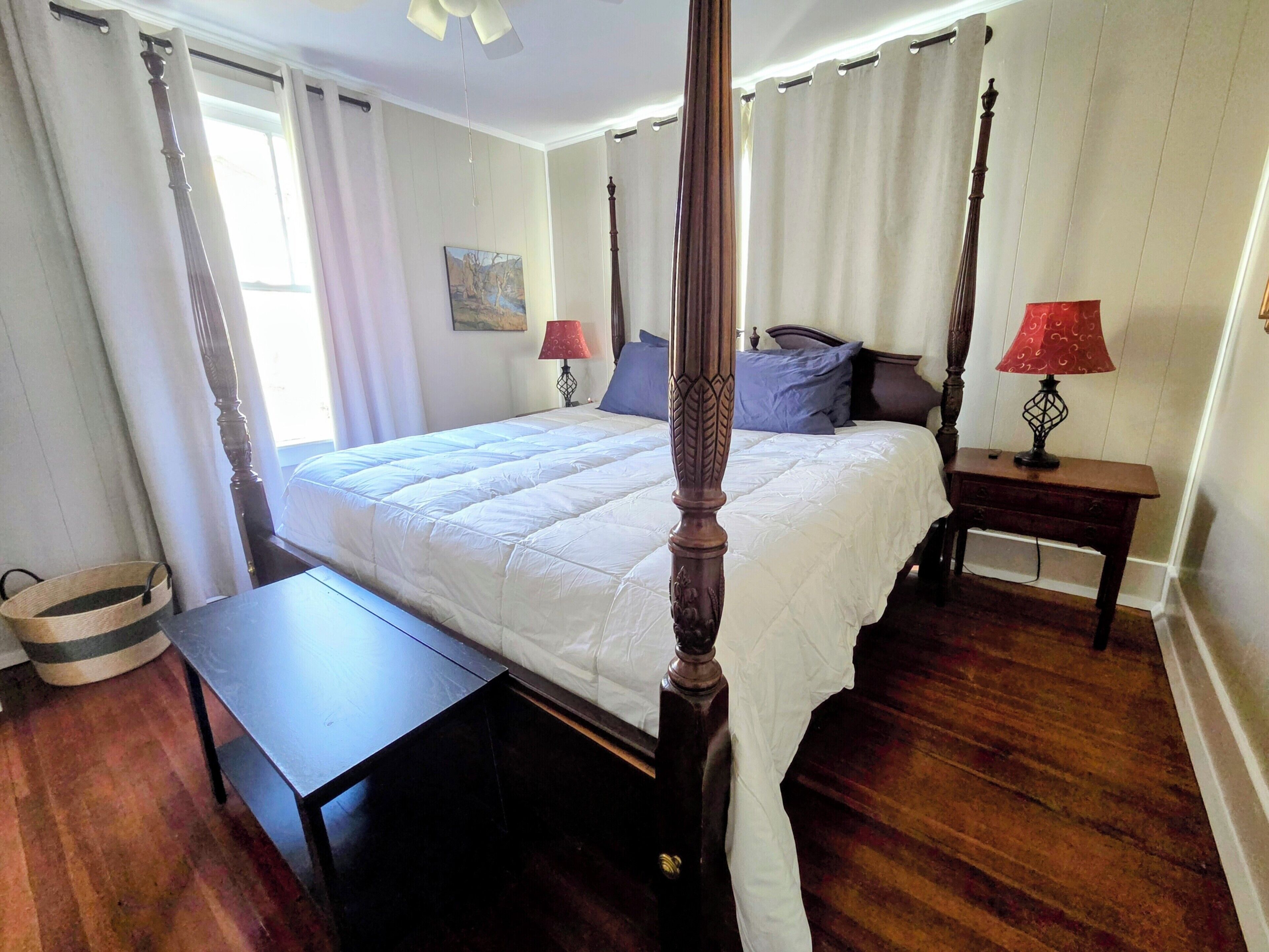 Een hemelbed met houten hoofdeinde, een nachtkastje met lamp en een schilderij aan de muur.