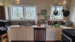 A modern kitchen with white cabinets, a stainless steel dishwasher, a gas stove, and a microwave. There are two windows providing natural light, and a variety of kitchen utensils and appliances are visible. - Historic Carrie Tucker Cottage with view of Spring River in charming Hardy, Ark. (Hardy)