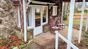 A stone building with a white door and windows, a covered porch with chairs, and a stone wall. - Historic Carrie Tucker Cottage with view of Spring River in charming Hardy, Ark. (Hardy)