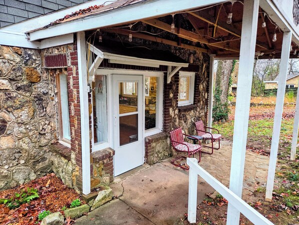 A stone building with a white door and windows, a covered porch with chairs, and a stone wall.