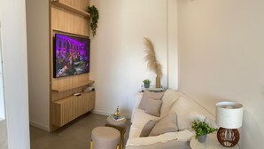 Un salon moderne avec un téléviseur à écran plat fixé à un mur en bois, un canapé beige, un petit pouf rond, une table d’appoint avec une lampe et une plante.