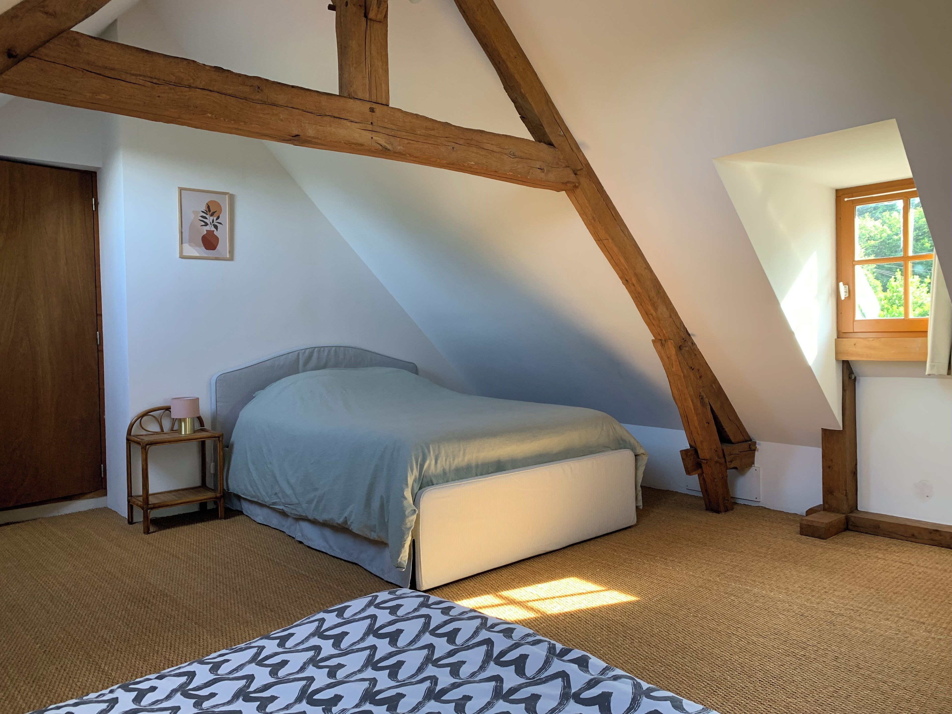 Une chambre avec un plafond en pente, un lit, une table de chevet avec une lampe et une fenêtre donnant sur un paysage verdoyant.