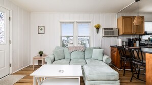 A modern living room with a sectional sofa, a small table, and a kitchen area with wooden cabinets and a dining set. - 2 Blocks to Beach | Downtown OC +  1 Parking Space (Ocean City)
