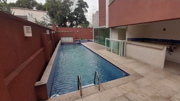 Una piscina exterior con plataforma de baldosas, barandilla de vidrio y extintor de incendios en la pared.