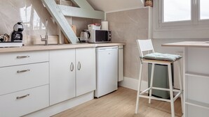 Une cuisine moderne avec des meubles blancs, un lave-vaisselle encastré, un four à micro-ondes et une bouilloire. On y trouve une chaise en bois avec un coussin et une petite plante en pot sur le rebord de la fenêtre.