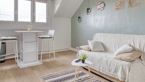 Un salon avec un plafond haut, un canapé blanc, une petite table avec une plante et une fenêtre avec des stores.
