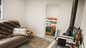 Un accogliente soggiorno con divano, un cane, un angolo cottura con lavello e utensili, e vista sulla camera da letto attraverso una porta aperta.