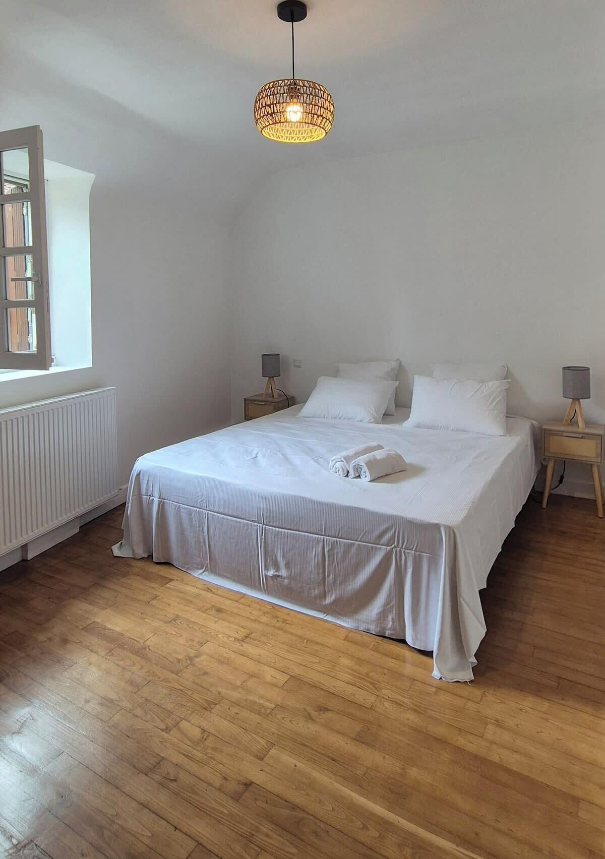Une chambre à coucher avec un grand lit, deux tables de chevet, un luminaire suspendu et une fenêtre avec des stores.