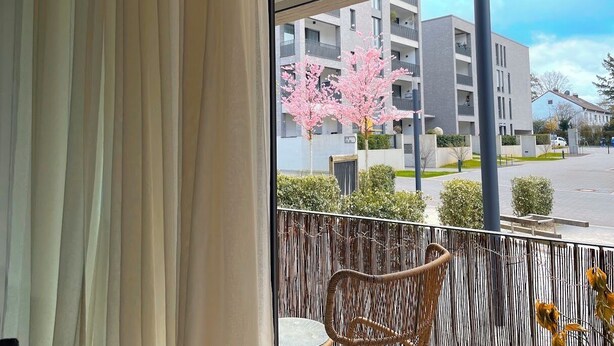A balcony with a wicker chair, a view of a modern building, and a cherry blossom tree. - Fabulous 1-bedroom apartment in Essen (Essen)