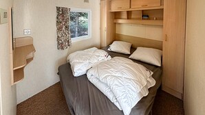 Een compacte slaapkamer met een bed, kussens, een raam met gordijnen en een houten kastje boven het bed.