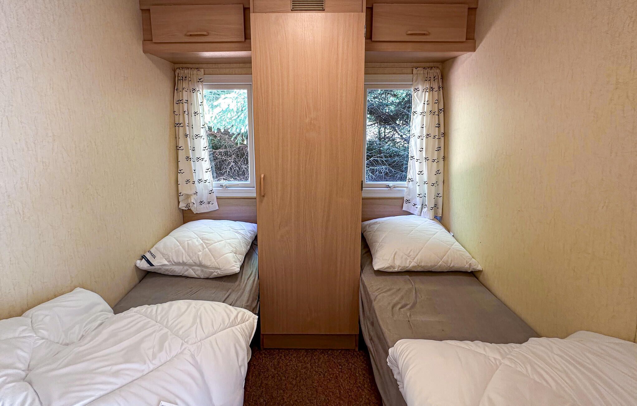 A small, narrow room with two single beds, each with a pillow and a blanket. There are two windows with curtains, and the room has a wooden ceiling with built-in lighting.