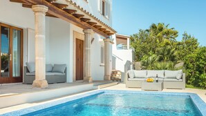 Un área junto a la piscina con una piscina azul cristalina, un sofá de mimbre y una pérgola de madera.