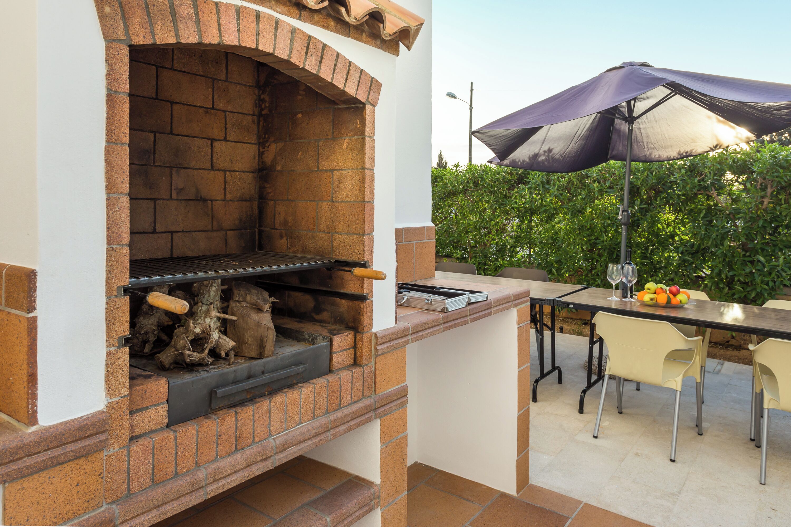 Une cuisine extérieure dotée d’un four en briques, d’une table à manger avec des chaises et d’un grand parasol pour créer de l’ombre.