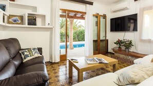 Una sala de estar con un sofá de cuero marrón, una mesa de centro de madera y un sofá blanco. Hay un televisor montado en la pared, una estantería con libros y objetos decorativos, y un ventanal corredero que da a un área exterior con piscina.