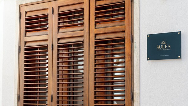 A wooden double door with horizontal slats, a plaque with the name 'SULEA', and a white wall with a textured lower half. - Suléa Horizon Apartment (Menfi)