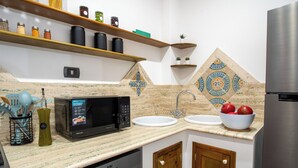 A kitchen with a microwave, sink, and a bowl of fruit. There are shelves above the counter with jars and a potted plant. - Suléa Horizon Apartment (Menfi)