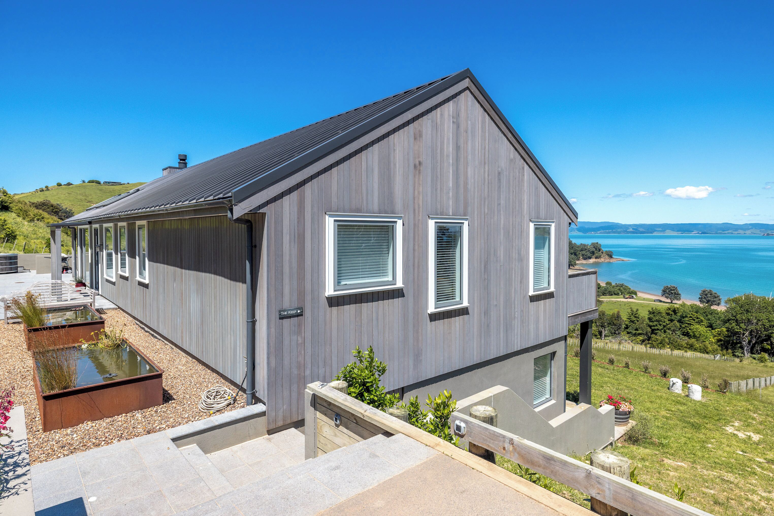 A modern two-story house with a grey exterior, a small deck, and a view of the sea.