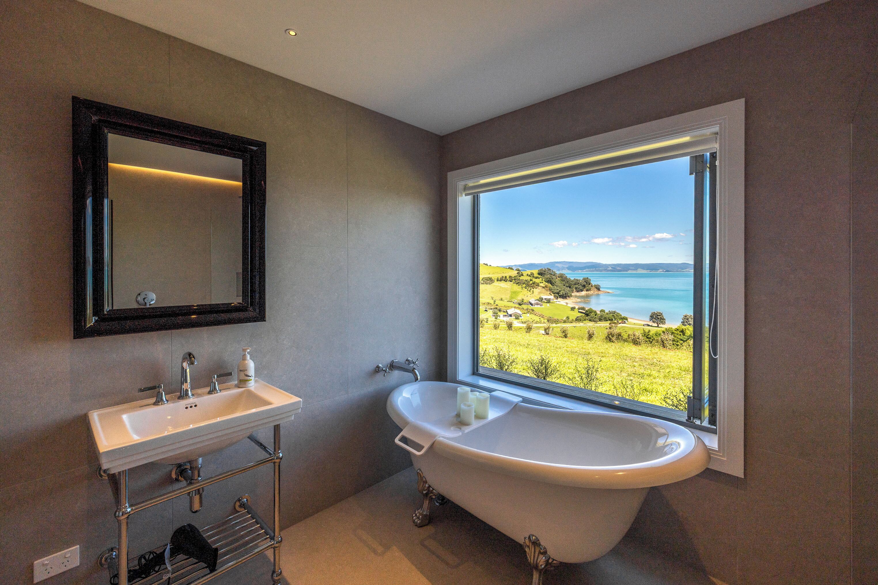 Un bagno con un'ampia finestra che offre vista su un paesaggio costiero, una vasca freestanding e un mobile lavabo con specchio e lavandino.