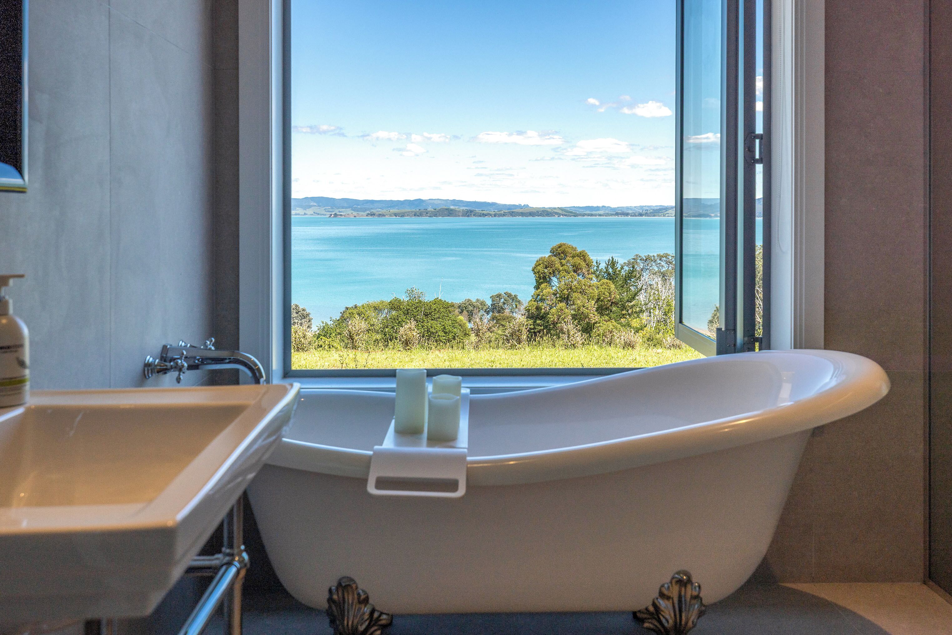 A bathroom with a freestanding bathtub, a sink, and a large window offering a view of a lake and distant hills.