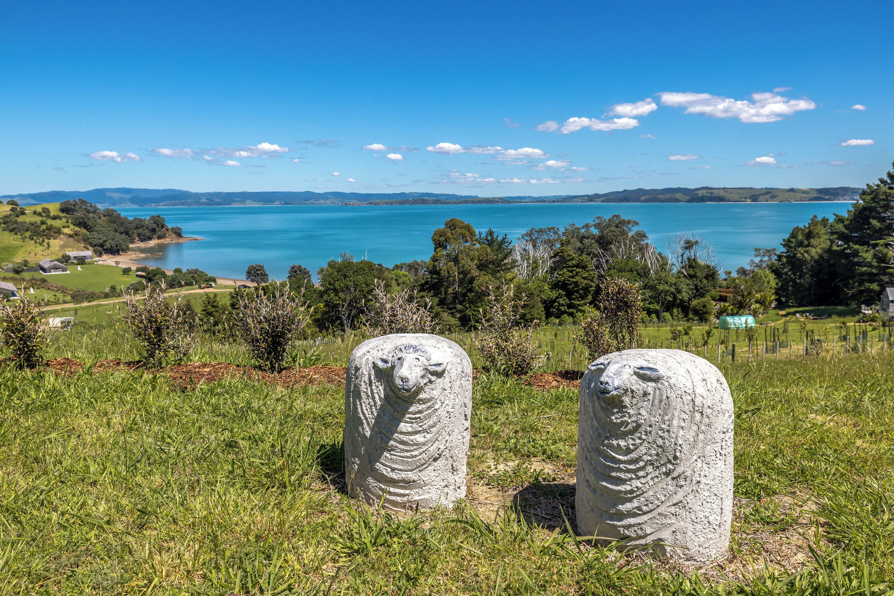 Deux statues de moutons en pierre, sur une colline herbeuse, d’où l’on aperçoit un lac et des collines au loin.
