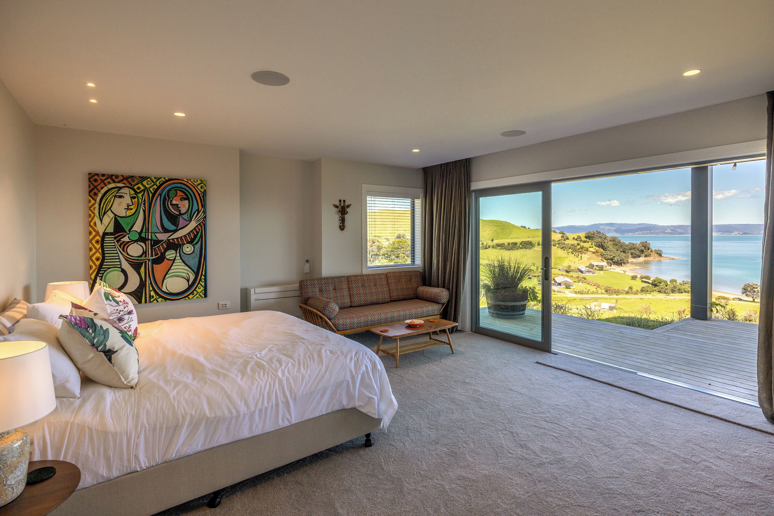 A bedroom with a large bed, a sofa, a painting, and a view of a lake.
