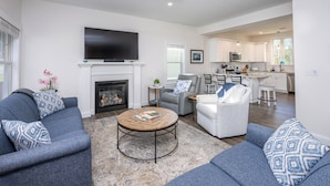 A modern living room with a fireplace, a television, and a kitchen in the background. - Resort style community, heated pools, club house, game room, 15 mins to beaches. (Arundel)