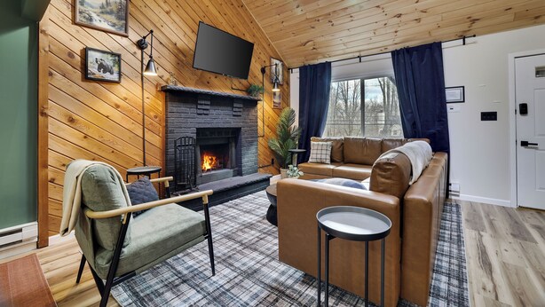 A cozy living room with a fireplace, wooden walls, a sofa, and a TV mounted on the wall. - NEW Lodge•HotTub.Game Room.Firepit.Theater.Hammocks (Tobyhanna)