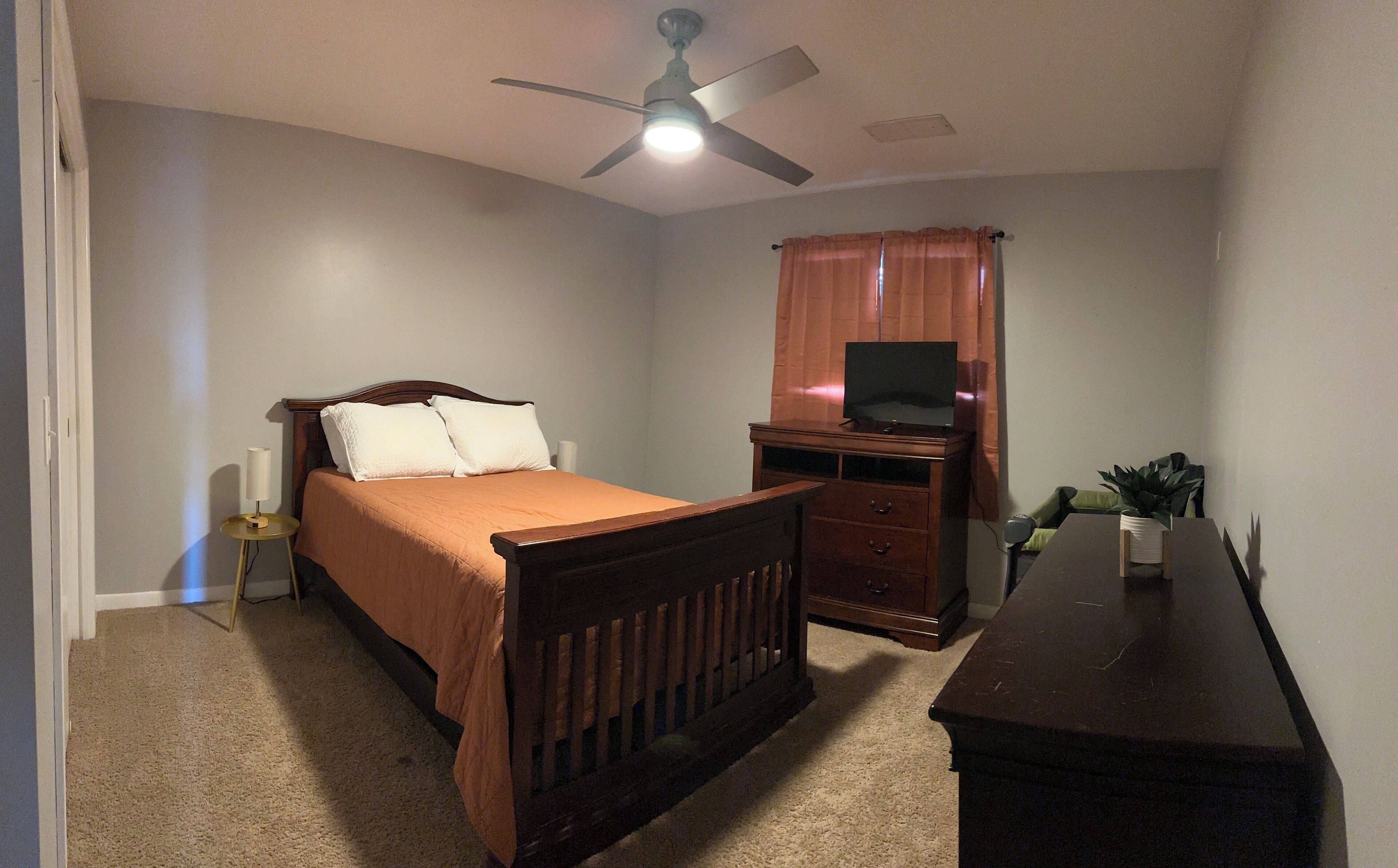 A bedroom with a bed, a TV on a stand, a desk, and a ceiling fan.