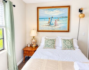 Een slaapkamer met een bed, een nachtkastje met een lamp, een schilderij aan de muur en een raam met gordijnen.