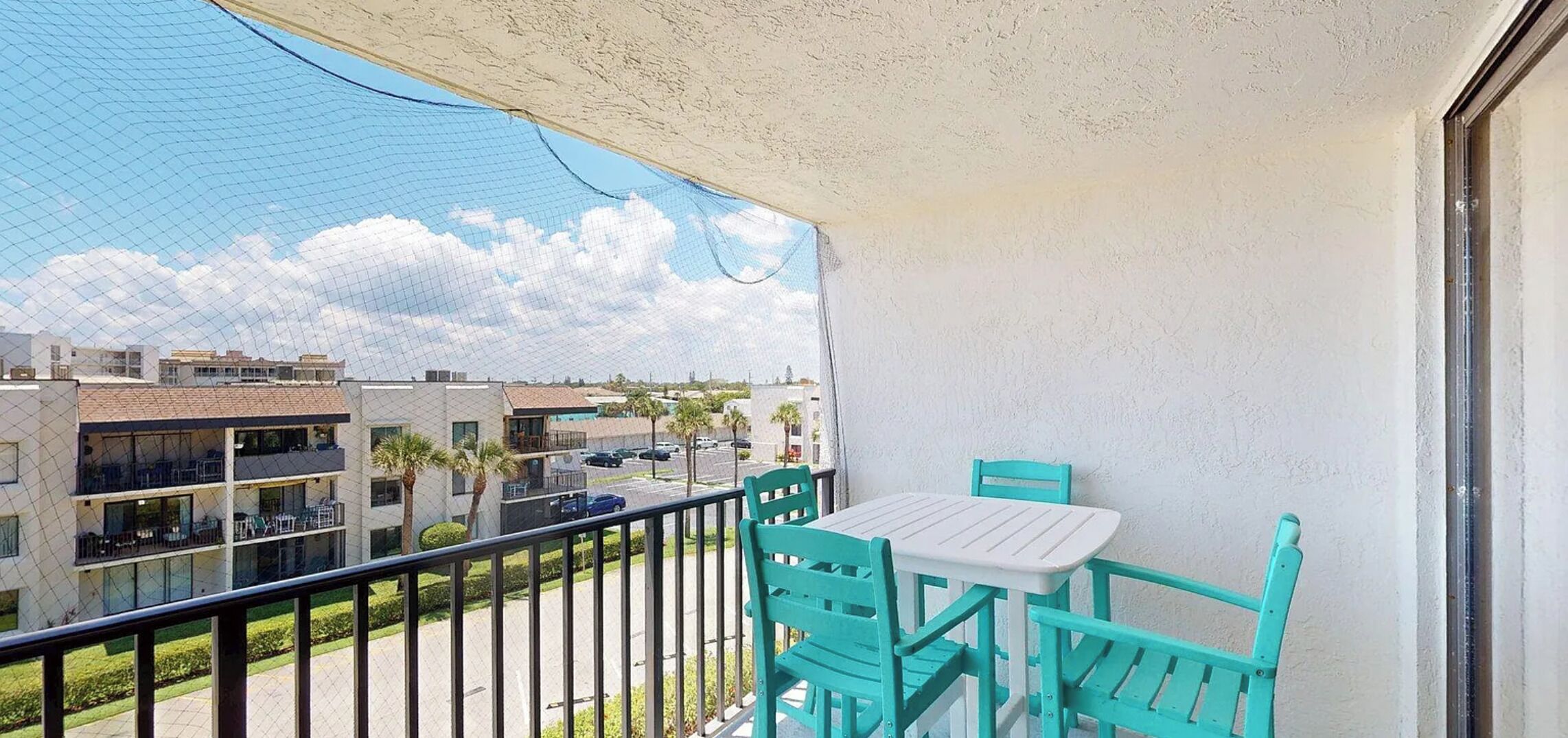 A balcony with turquoise chairs and a table, overlooking a residential area with buildings and parked cars.