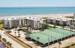 Un hotel de varios pisos con cancha de tenis, estacionamiento y vista al mar.