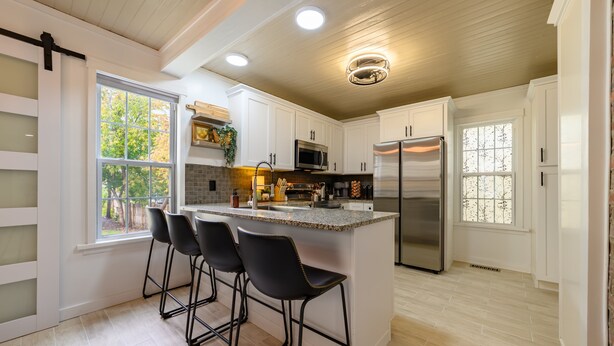 A modern kitchen with white cabinets, a stainless steel refrigerator, and a bar area with black stools. - Newly Renovated | Old Town | King Suite | Fire Pit | Garage (Alpena)