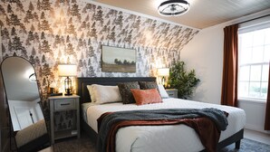 A bedroom with a large bed, two bedside tables with lamps, a mirror, a painting on the wall, and a window with curtains. - Newly Renovated | Old Town | King Suite | Fire Pit | Garage (Alpena)