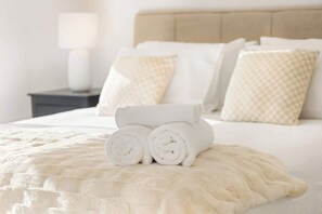 Un letto rifatto con asciugamani bianchi arrotolati, una testiera beige e cuscini coordinati.
