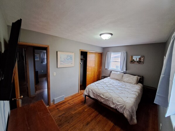 Ein Schlafzimmer mit Bett, Deckenleuchte, Fenster mit Vorhängen und einer Tür zu einem anderen Raum.
