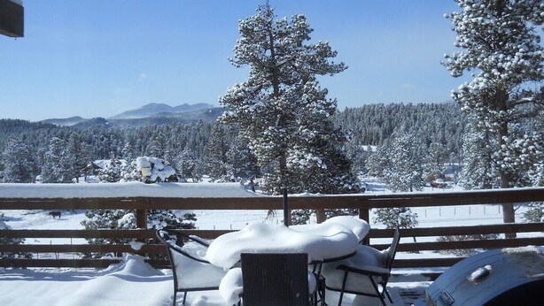 Een houten terras met sneeuw bedekte stoelen en een barbecue, met uitzicht op een besneeerd landschap met bomen en bergen.