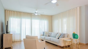 Un salon comprenant un canapé, un fauteuil, deux petites tables d’appoint, un ventilateur de plafond et un téléviseur posé sur un meuble.