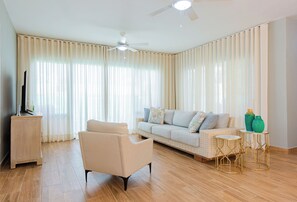 Ein Wohnzimmer mit einer Couch, einem Sessel, zwei kleinen Beistelltischen, einem Deckenventilator und einem Fernseher auf einem Schrank.
